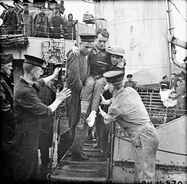 Black and white photograph. A sailor and soldier carry a wounded soldier down the gangplank of a large boat where two more sailors wait to take him. Men watch from the upper decks of the boat.
